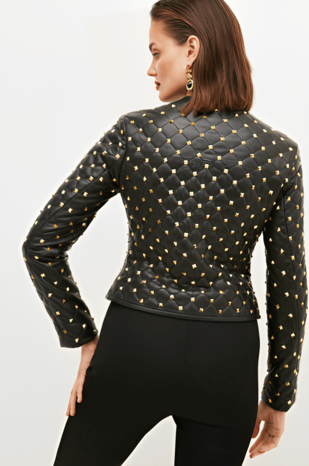 A woman shown from the back wearing a black quilted jacket covered in gold studs, paired with black pants, against a plain light background.
