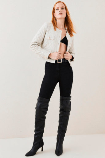 A model with red hair stands against a plain off-white studio background wearing a light beige quilted jacket, black cropped bralette, black skinny jeans, and black over-the-knee heeled boots.