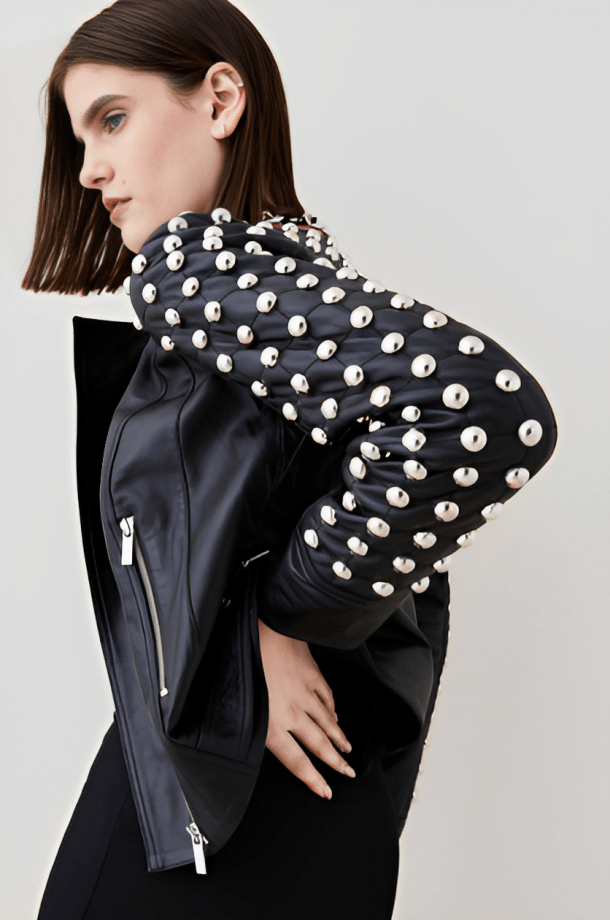 A model wearing a black leather jacket with dramatic, oversized sleeves heavily embellished with large silver dome studs, photographed against a light gray studio background.