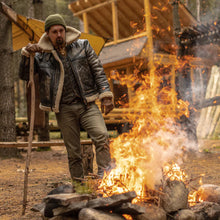 Man in a leather jacket and green beanie standing near a campfire with a cabin in the background.