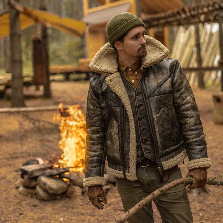 Man in a leather jacket with fur lining standing near a campfire in a rustic setting