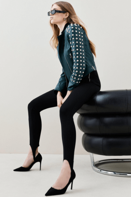 A woman wearing a dark teal studded leather jacket, black skinny pants, and black pointed-toe heels sits on a black leather chair against a light beige studio background.