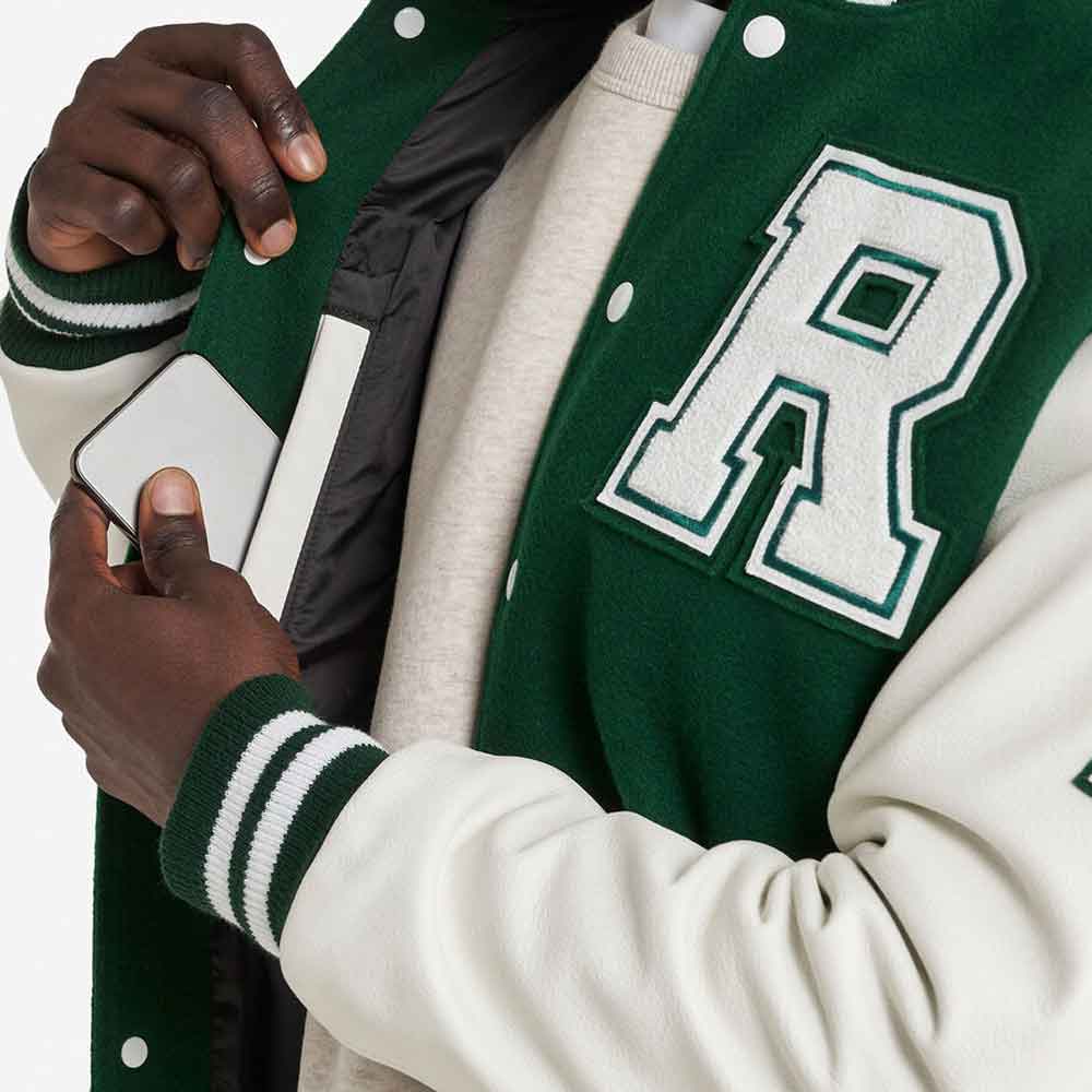 A close-up of a person wearing a green and off-white varsity jacket with a large white letter 'R' on the chest, holding a white phone inside the jacket pocket.