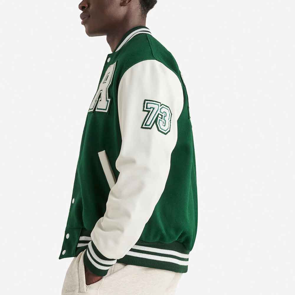A side-profile view of a person wearing a green and off-white varsity jacket with '73' embroidered on the sleeve and a large letter on the chest, paired with light beige pants against a plain white background.