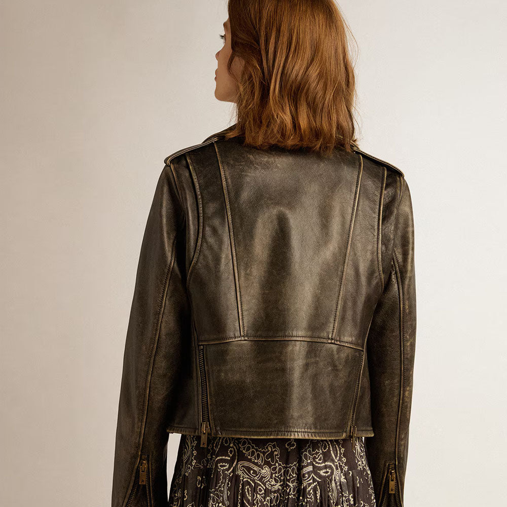 A woman viewed from the back wearing a cropped distressed leather jacket in a dark brownish-gray color, paired with a patterned skirt, against a plain light background.