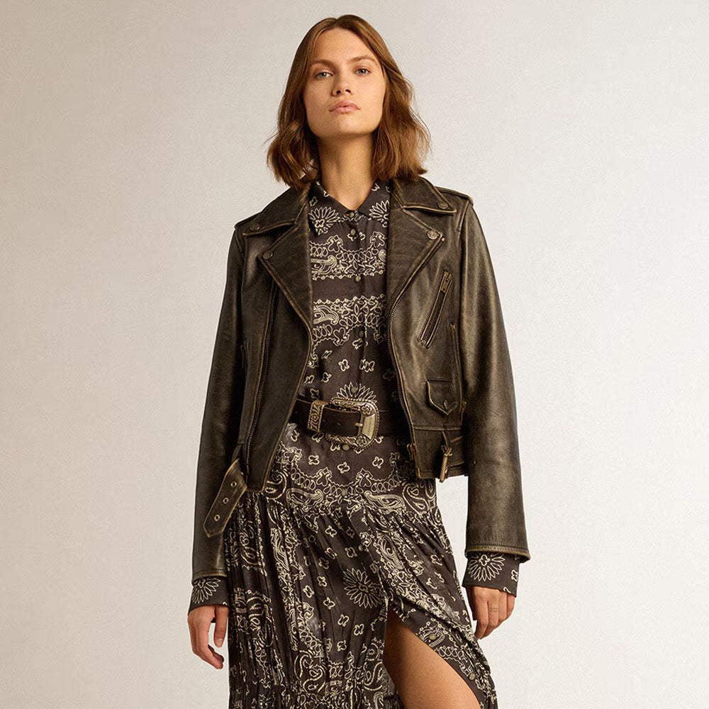 A woman models a dark brown distressed leather moto jacket over a brown and beige paisley print dress, set against a plain light gray studio background.