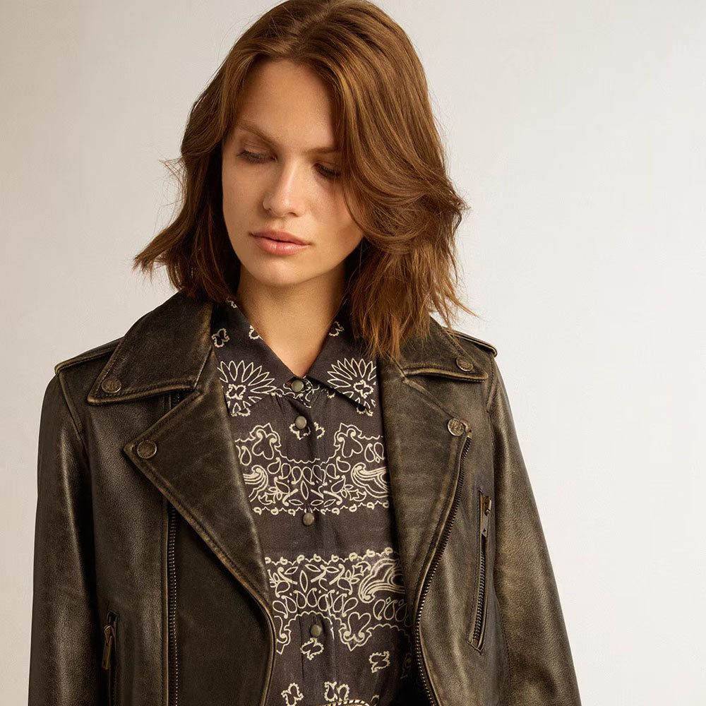A woman wearing a distressed dark brown leather moto jacket over a dark gray bandana print button-up shirt, photographed against a neutral light gray background.