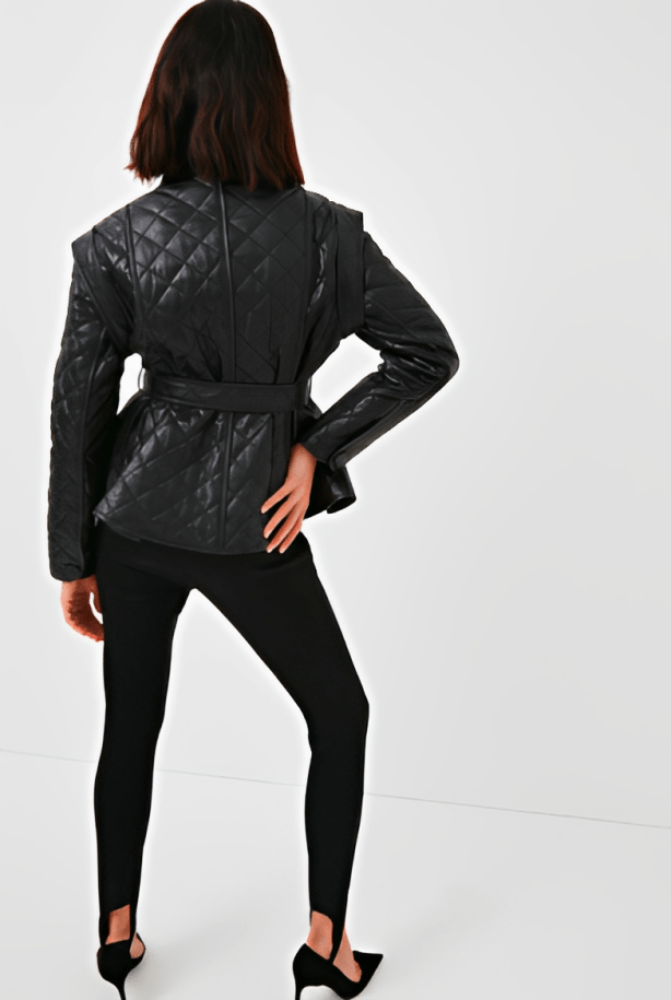 A woman shown from the back wearing a black quilted belted jacket, black leggings, and black heels against a plain light gray studio background.