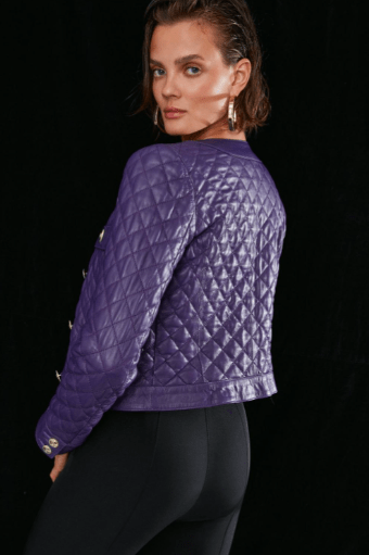 A woman wearing a purple quilted cropped jacket with gold-tone buttons, photographed against a solid black background.