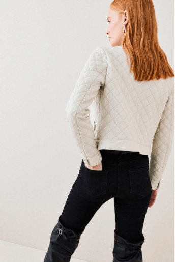 A woman with red hair is shown from behind wearing a cream-colored quilted cropped jacket and black pants against a plain white studio background.