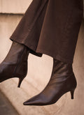 Dark brown pointed-toe ankle boots with kitten heels, worn with matching dark brown wide-leg jeans against a neutral concrete wall.