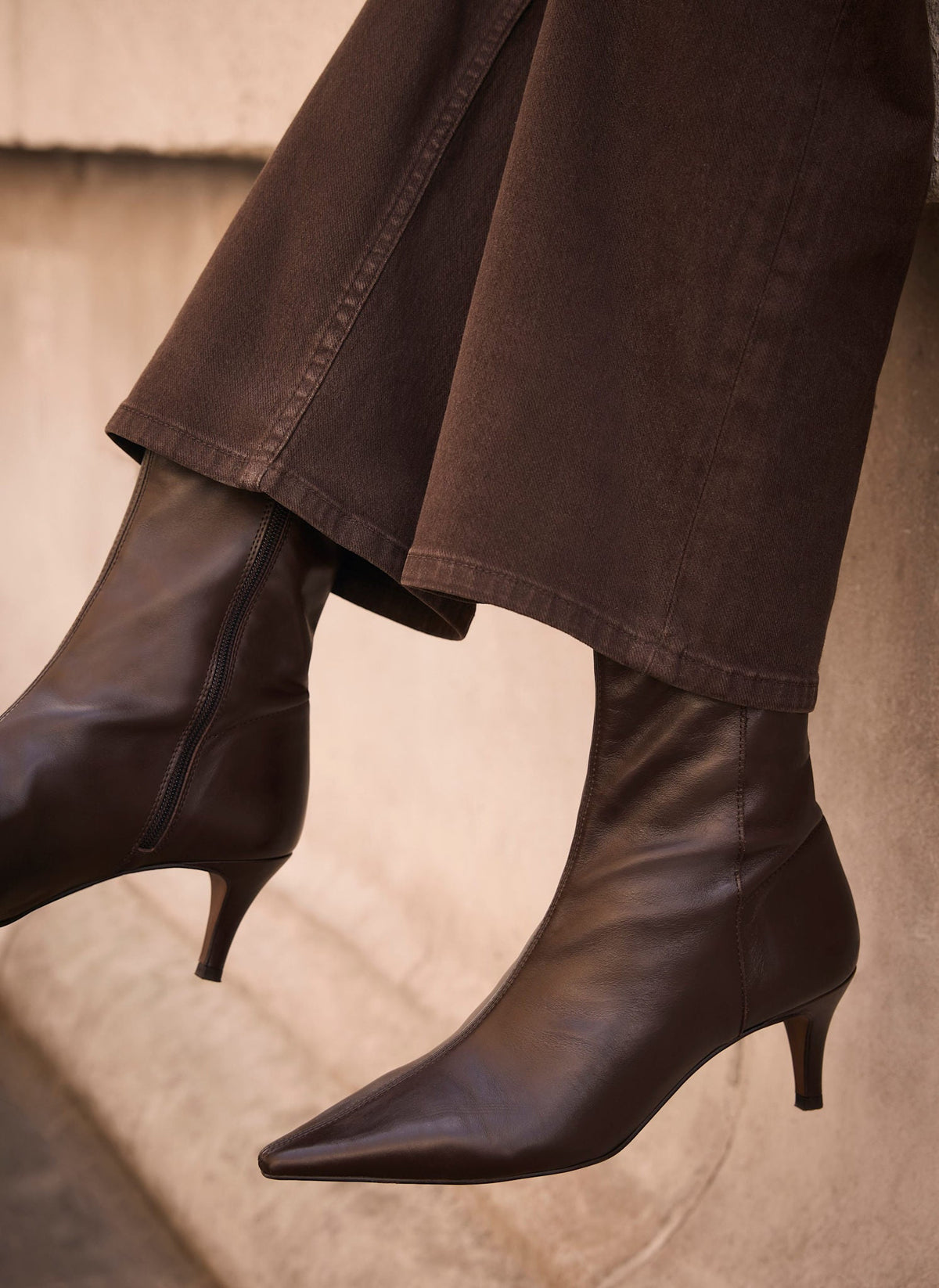 Dark brown pointed-toe ankle boots with kitten heels, worn with matching dark brown wide-leg jeans against a neutral concrete wall.