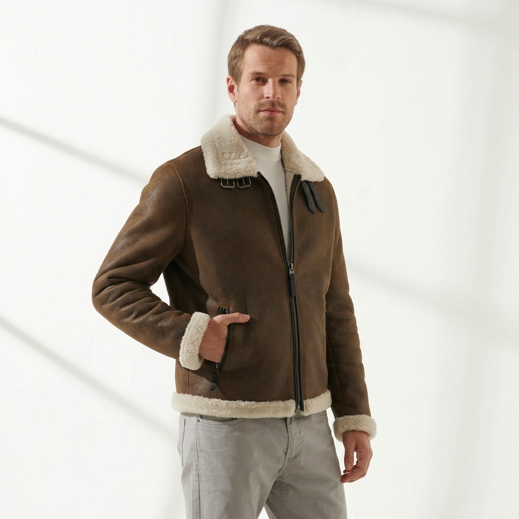 A man wearing a brown shearling-lined aviator jacket with a cream-colored collar and cuffs, paired with light gray trousers, standing against a bright white background with soft diagonal shadows.
