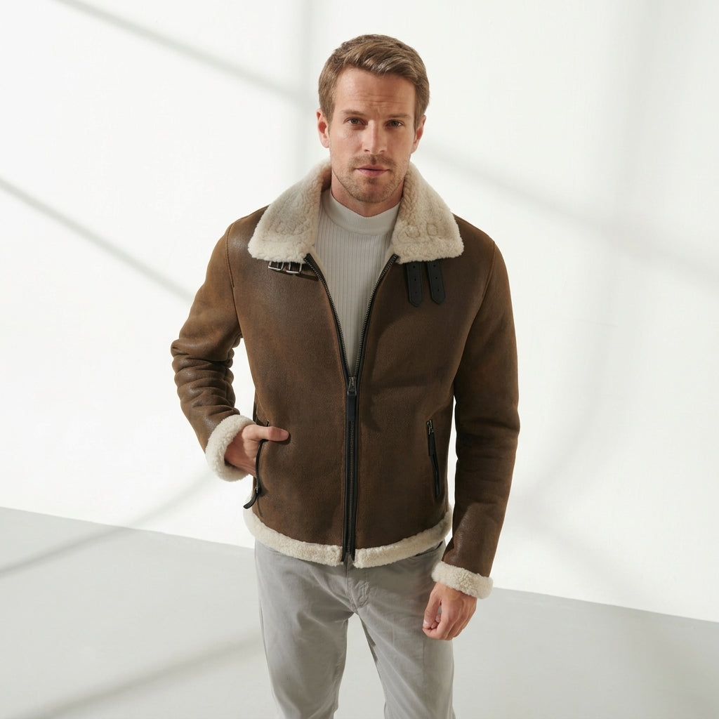 A man wearing a brown shearling-lined aviator jacket over a light gray ribbed sweater and light gray trousers, standing against a white wall with soft diagonal shadows.