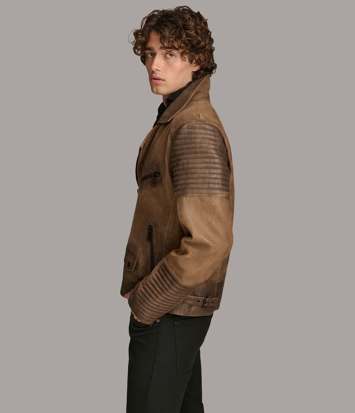 A man wearing a brown leather motorcycle jacket with textured horizontal paneling on the sleeves, shown in profile against a solid gray background.