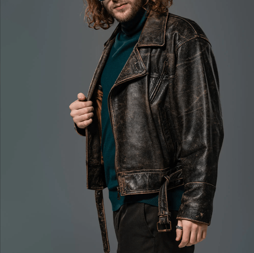 A man wearing a distressed, dark brown leather motorcycle jacket over a dark teal turtleneck sweater and black pants, shown from the waist up against a solid dark gray studio background.