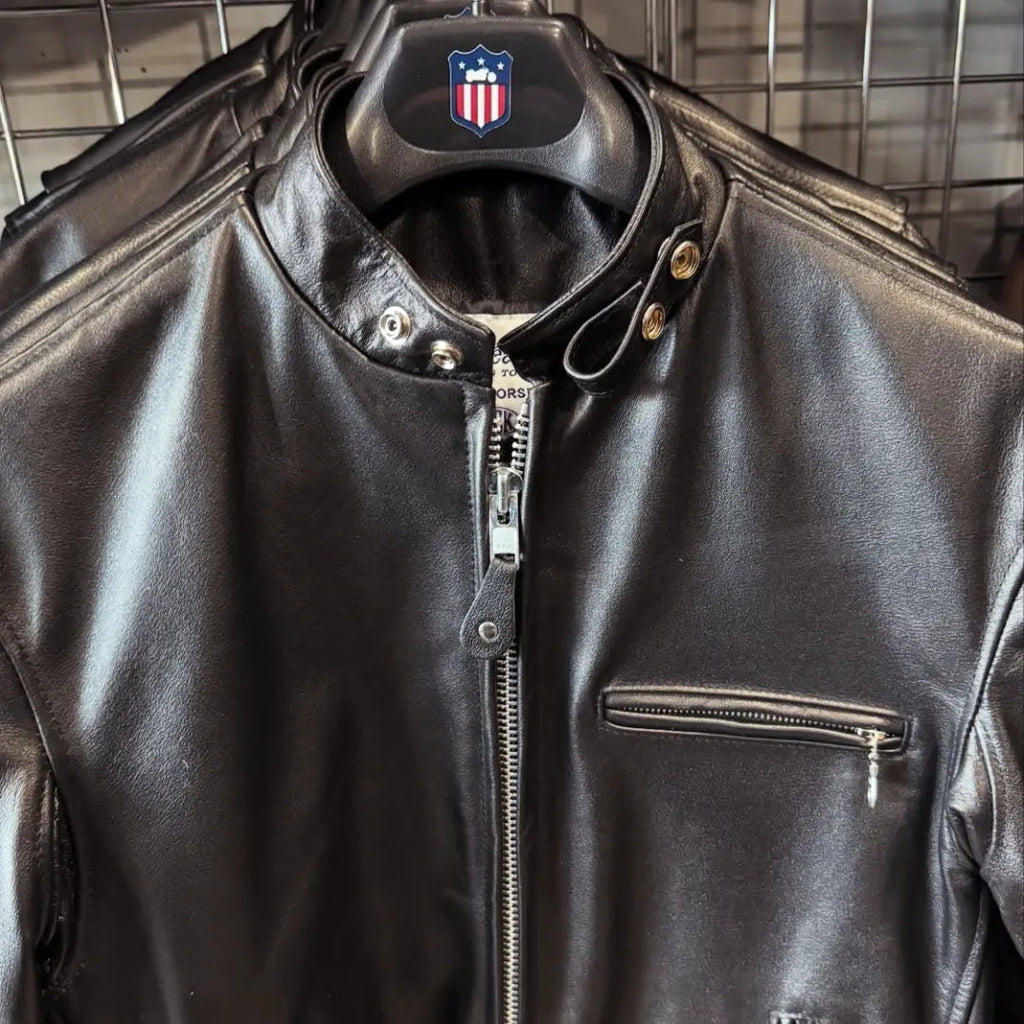 Black leather motorcycle jacket with a silver zipper, snap-button collar, and zippered chest pocket, shown hanging on a black hanger with a shield logo.