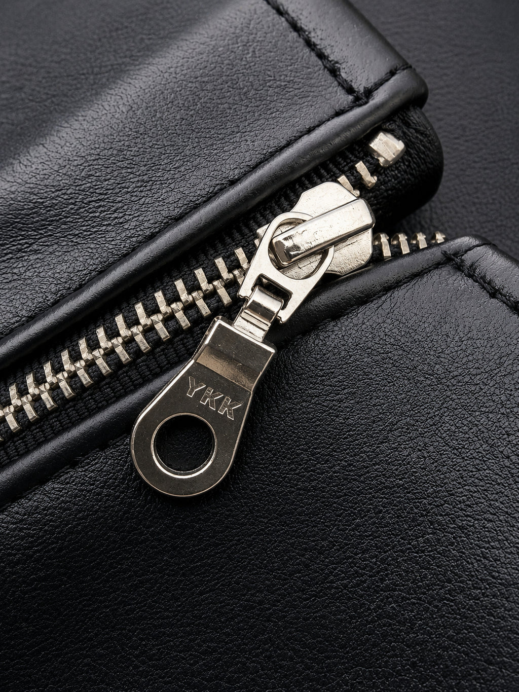 Close-up of a black leather zipper pull with silver metal teeth and a circular keyhole-shaped tab engraved with the brand name YKK.