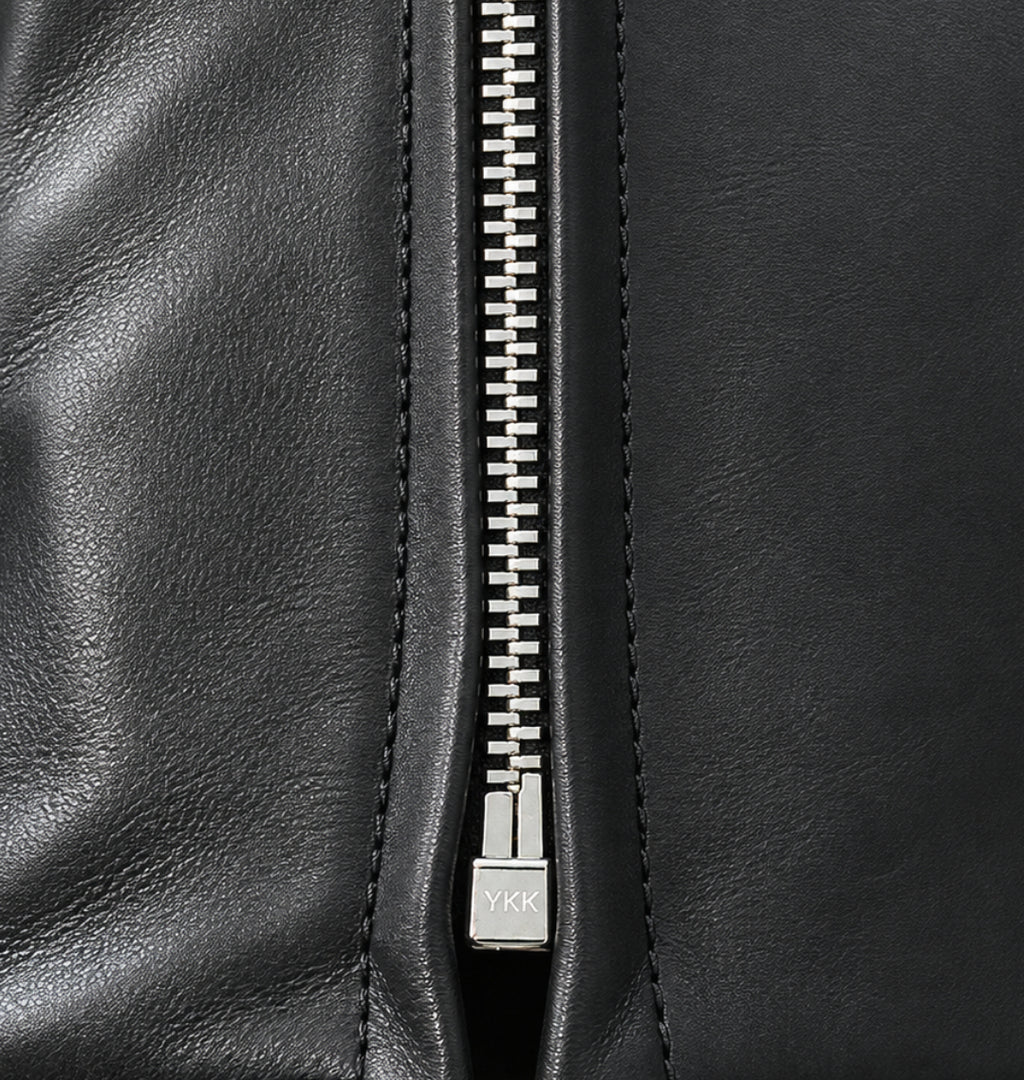 Close-up of a black leather zippered pouch or clutch, showing the texture of the leather, the silver metal zipper, and the pull tab on a white background.