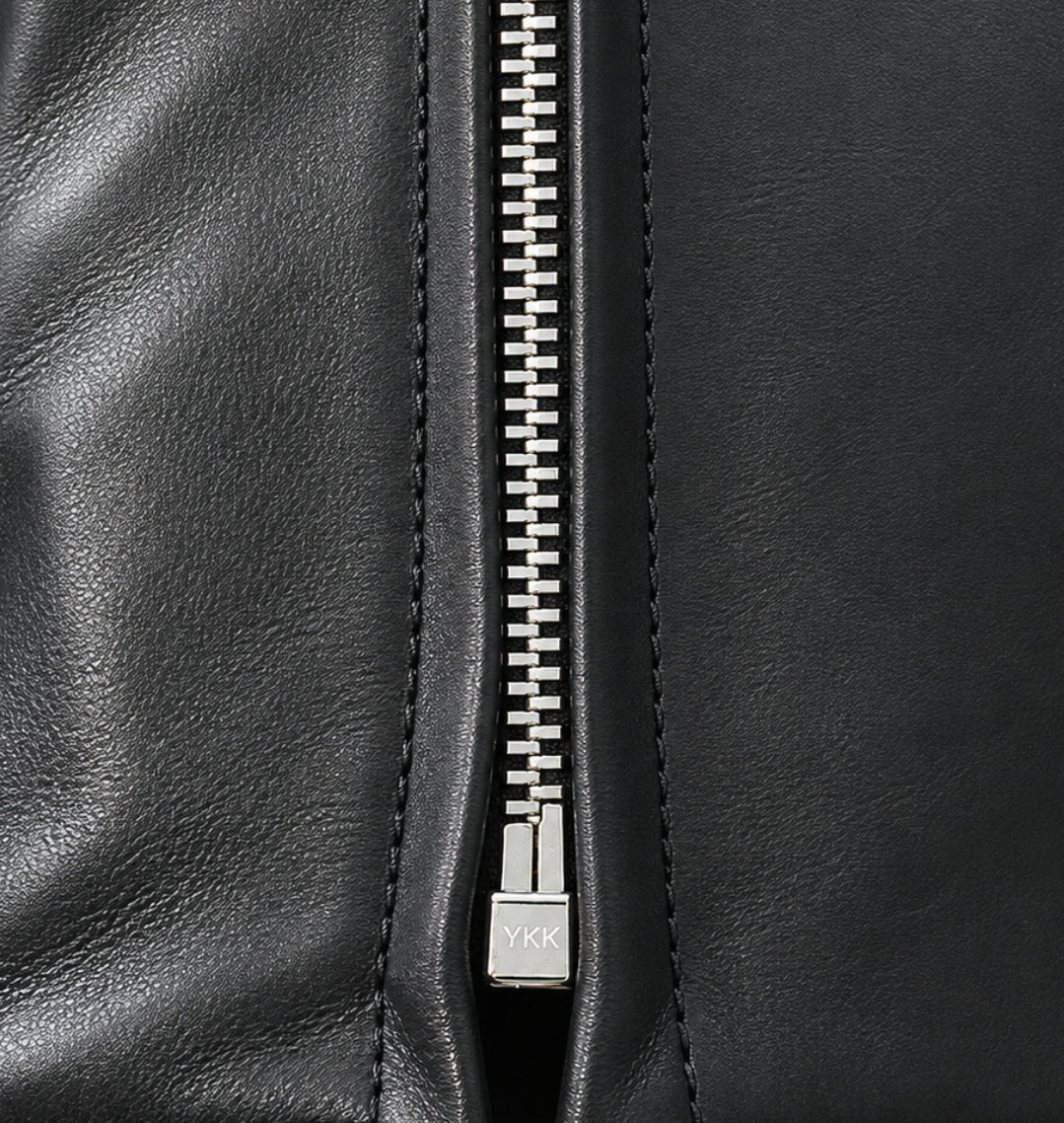 Close-up of a black leather zippered pouch or clutch, showing the texture of the leather, the silver metal zipper, and the pull tab on a white background.