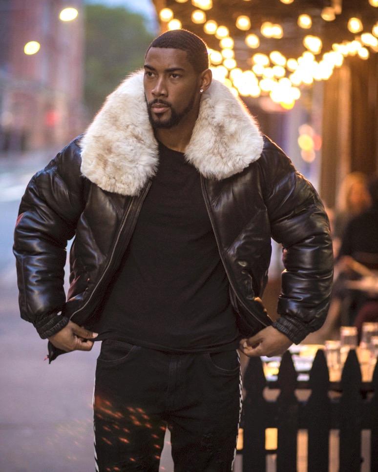 Man wearing a black leather jacket with a white fur collar in an urban setting.