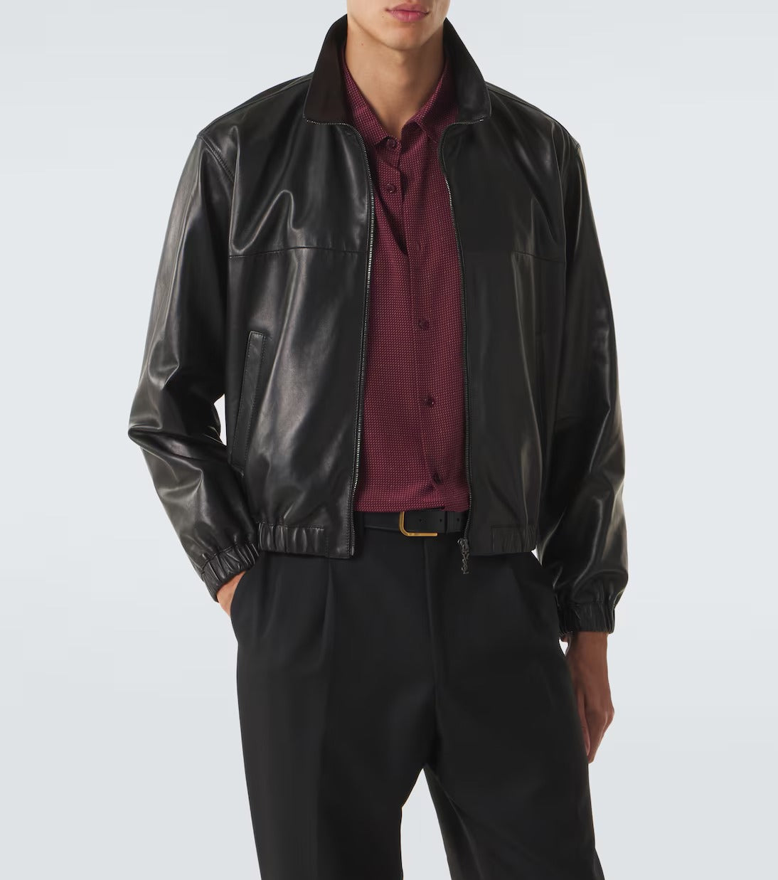 A man wearing a black faux-leather bomber jacket over a burgundy button-down shirt and black trousers, photographed against a light gray studio background.