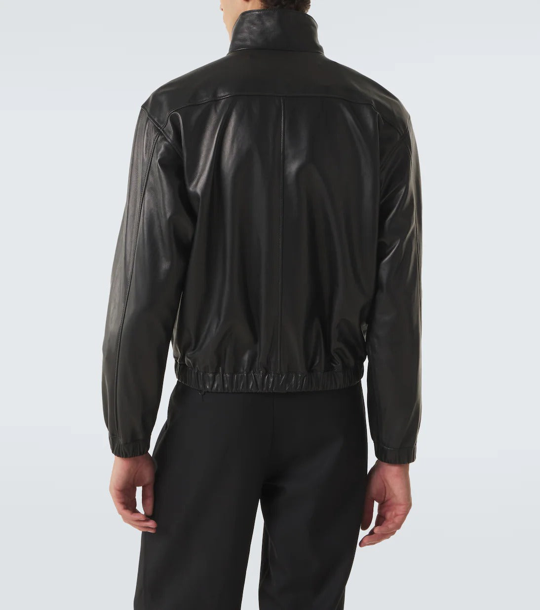 A person shown from the back wearing a black leather jacket with a high collar and elastic hem, paired with black pants, against a light gray studio background.