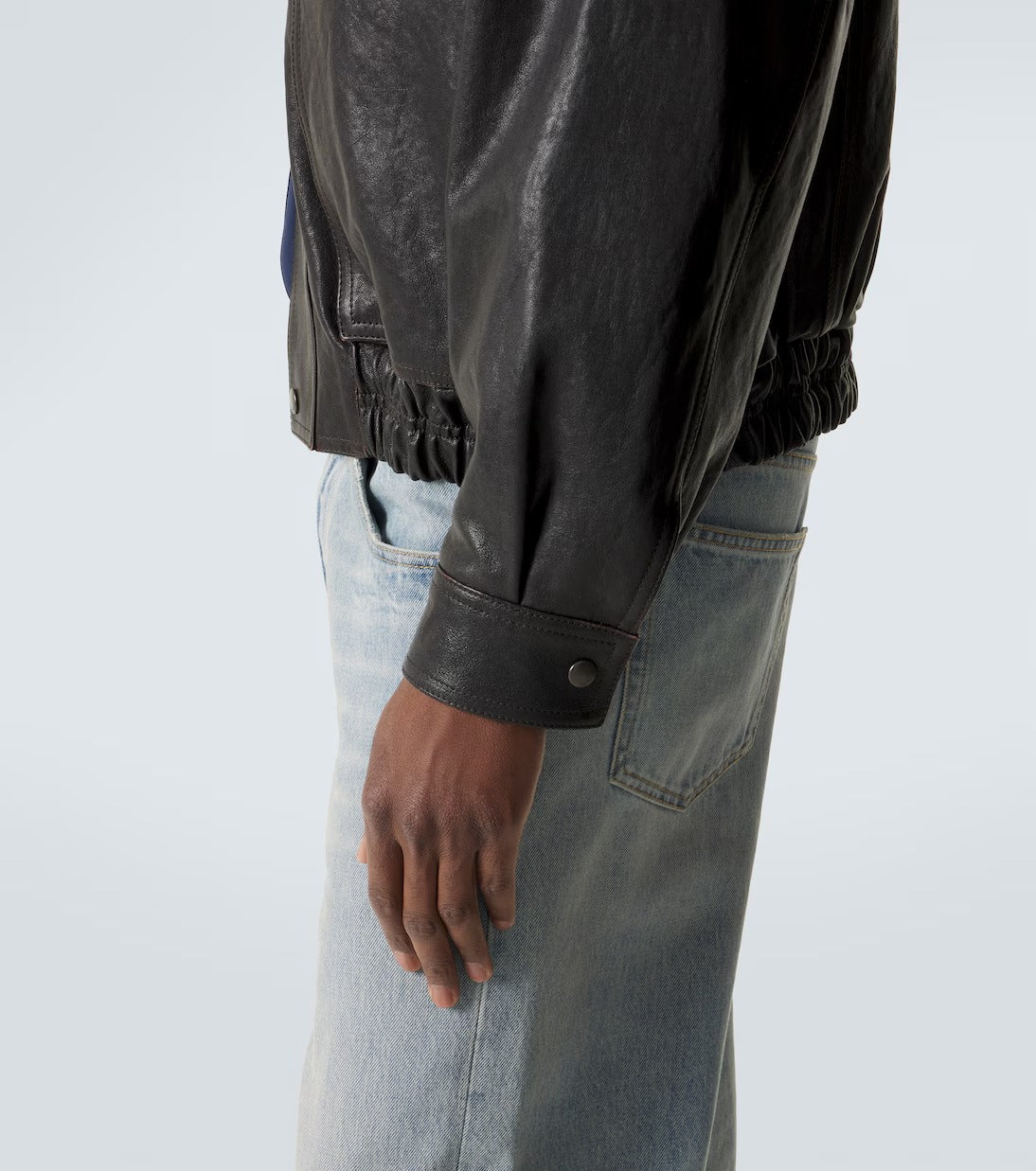 Close-up side view of a person wearing a black leather jacket with a gathered elastic hem and a buttoned cuff, paired with light-wash blue jeans against a clean light gray studio background.