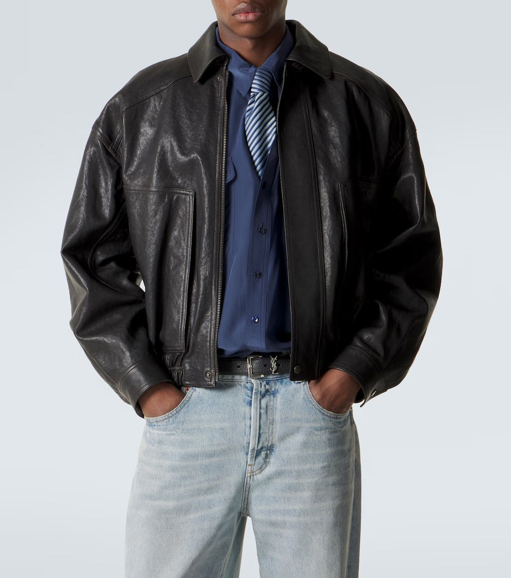 A man wearing a black leather jacket over a blue button-down shirt with a striped tie and light-wash jeans, photographed against a light gray studio background.