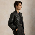 A woman in profile wearing a black quilted leather bomber jacket over a black top and dark trousers, standing against a warm beige textured studio background.