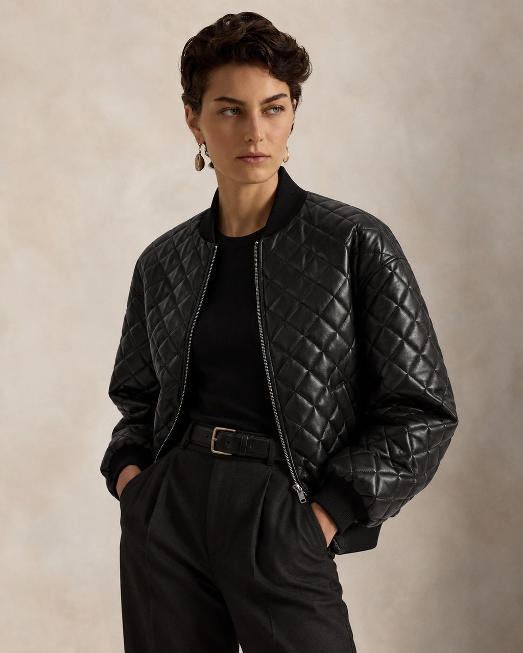 A model wearing a black quilted bomber jacket over a black top and black trousers, standing against a textured beige studio background.