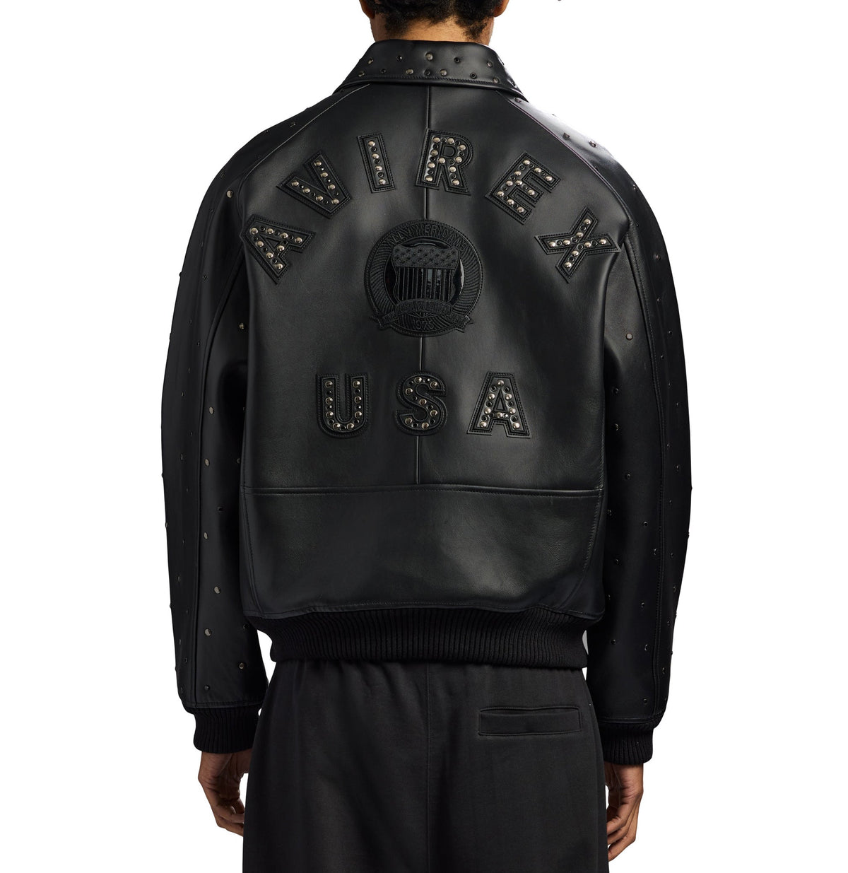 A black leather bomber jacket shown from the back on a model, featuring 'AVIREX USA' lettering and a central crest patch embellished with silver studs, set against a plain white background.