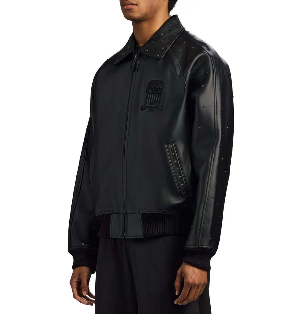 A man wearing a black leather bomber jacket with studded details and a tonal embroidered emblem on the chest, shown against a plain white background.