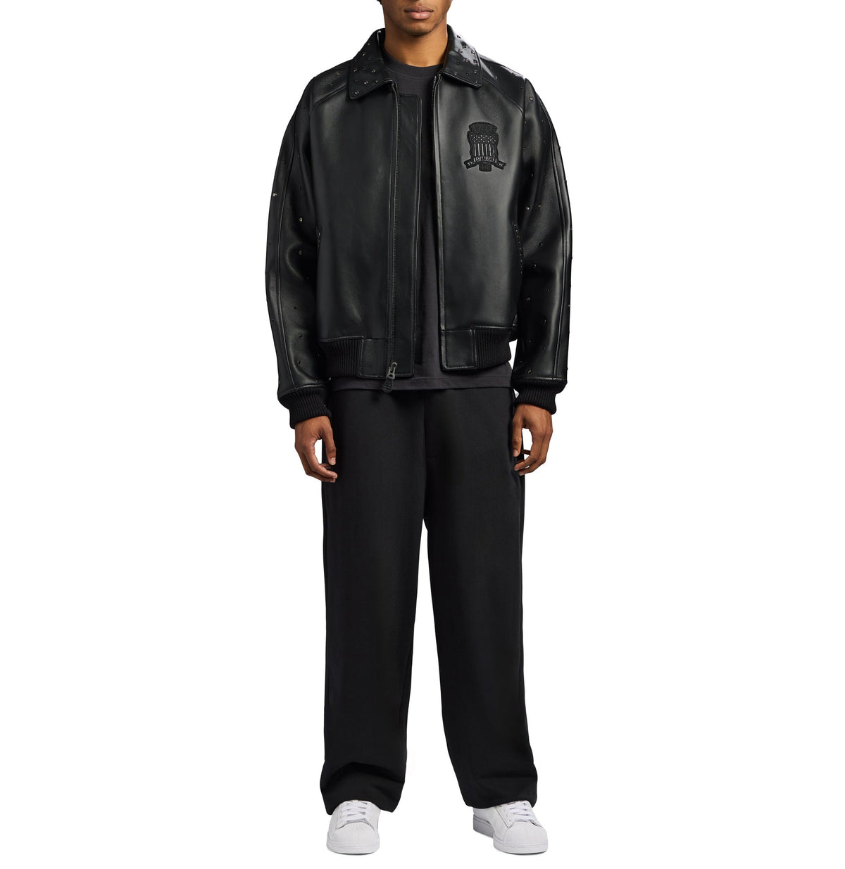 A model wearing a black leather bomber jacket with a tonal embroidered logo on the left chest, paired with black sweatpants and white sneakers, shot against a solid white background.