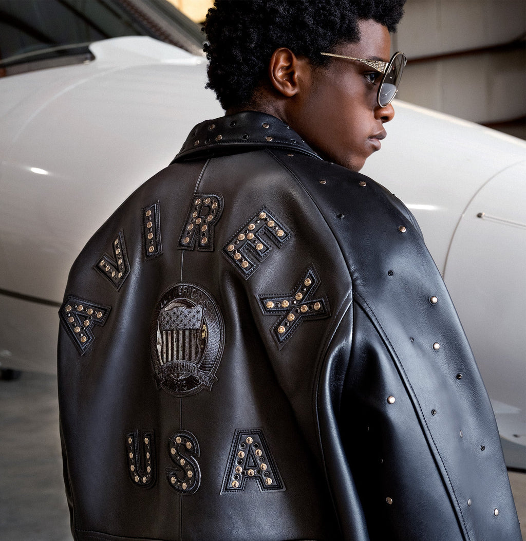 A person wearing a black leather jacket with large studded lettering on the back reading 'AVIREX USA' and a central crest patch, photographed in front of a white airplane in a hangar.