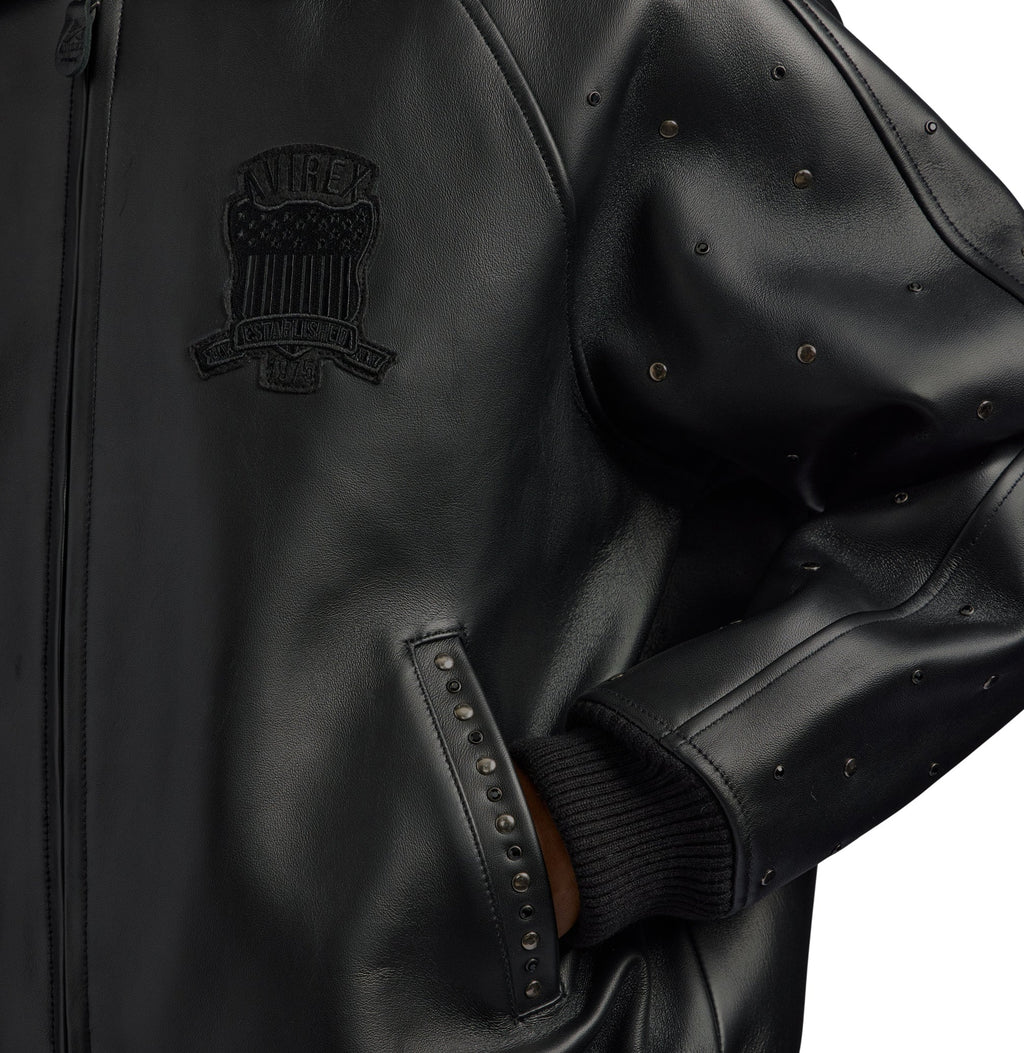 Close-up of a black leather jacket featuring a tonal black embroidered crest patch on the chest, studded sleeve details, and a ribbed black knit cuff.