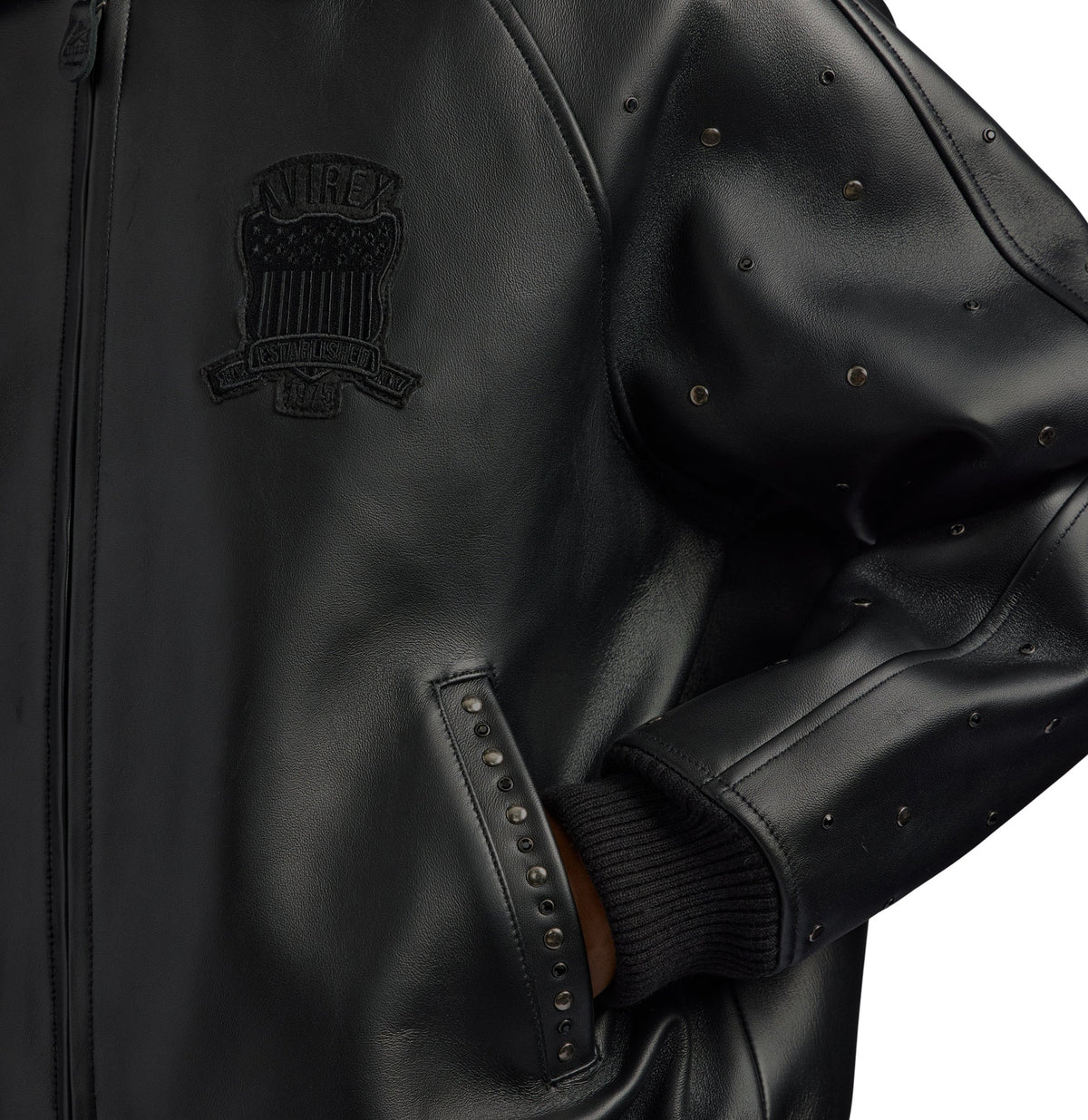 Close-up of a black leather jacket featuring a tonal black embroidered crest patch on the chest, studded sleeve details, and a ribbed black knit cuff.