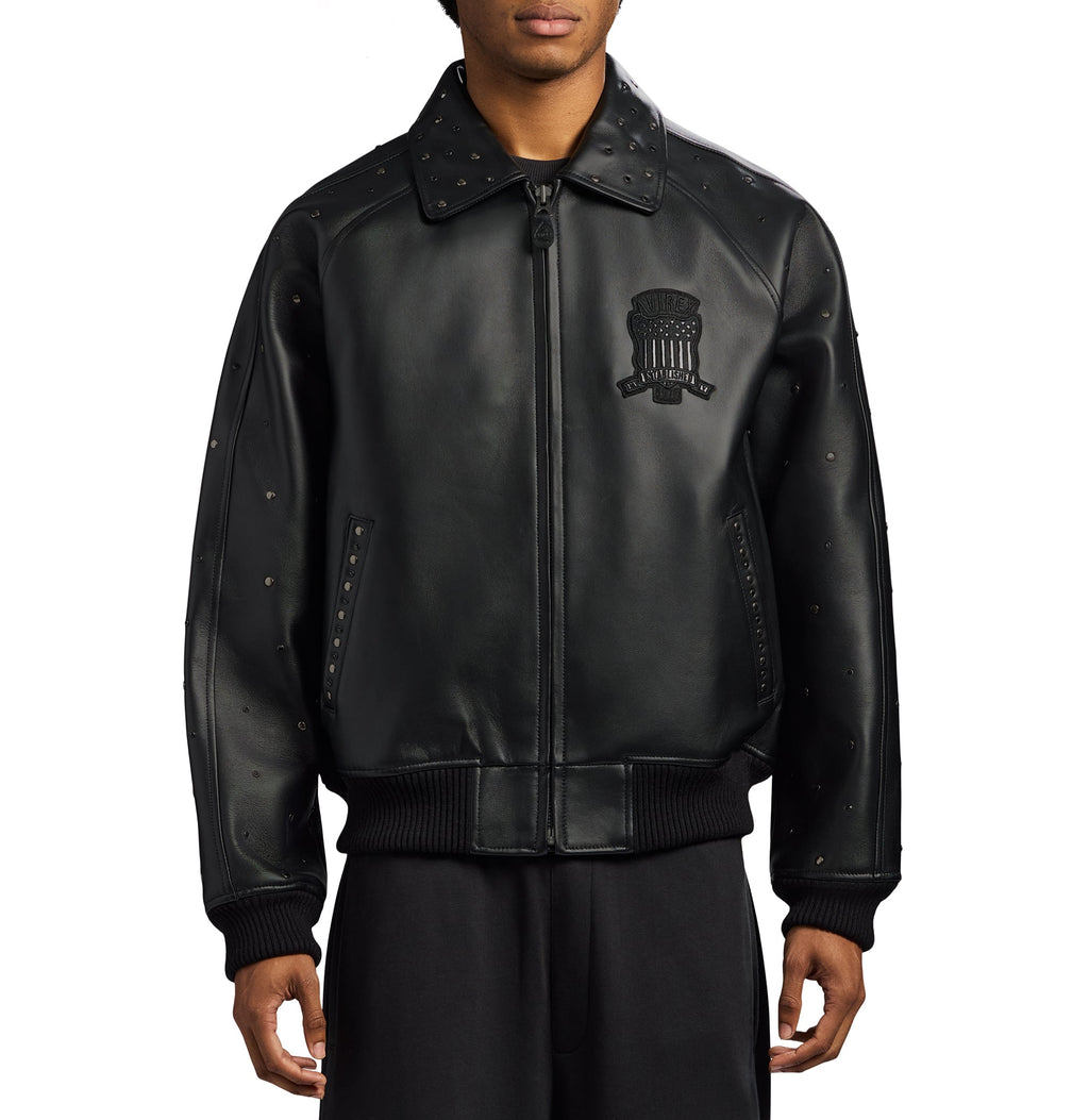 A black leather bomber jacket with silver-tone studs and a black embroidered crest on the left chest, worn by a model against a plain white background.