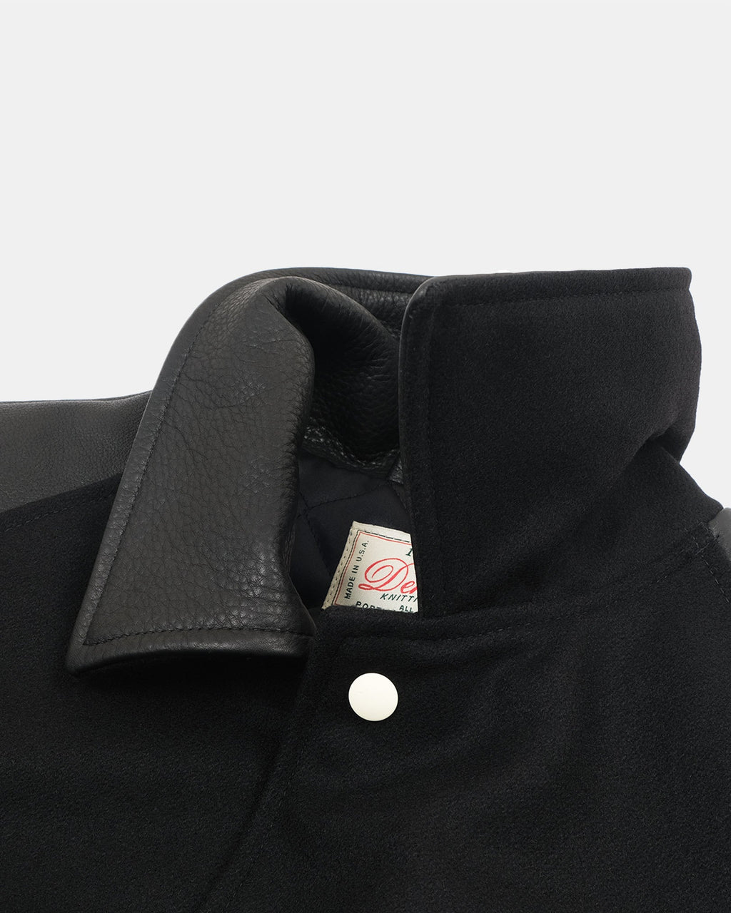 Close-up of a black jacket collar showing a textured black leather lapel, a white snap button, and a small interior brand tag.