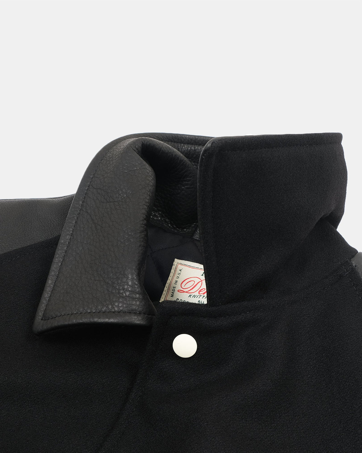 Close-up of a black jacket collar showing a textured black leather lapel, a white snap button, and a small interior brand tag.