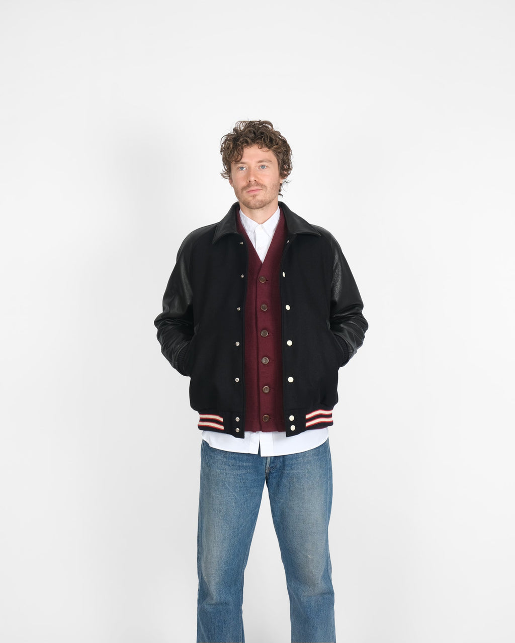 Man wearing a black varsity jacket with red and white accents, blue jeans, and a white shirt on a white background.