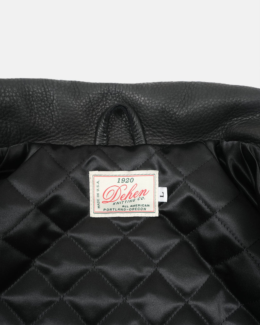 Close-up of a black quilted jacket or coat interior with a visible sewn-in label reading 'Deher Printing Co.' and 'Portland, Oregon' on a light gray background.
