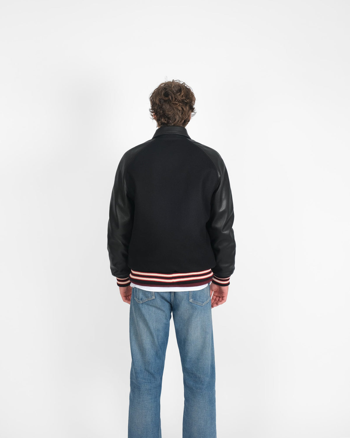 Person wearing a black varsity jacket with leather sleeves and blue jeans on a white background