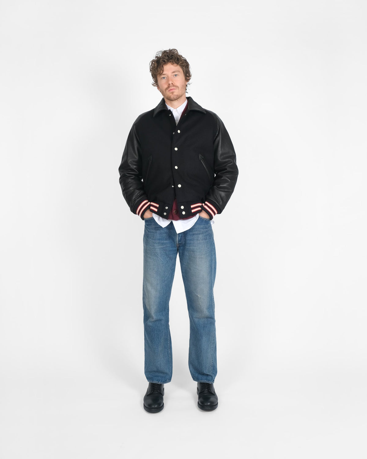 Man wearing a black leather jacket with red and white accents, blue jeans, and black shoes on a white background