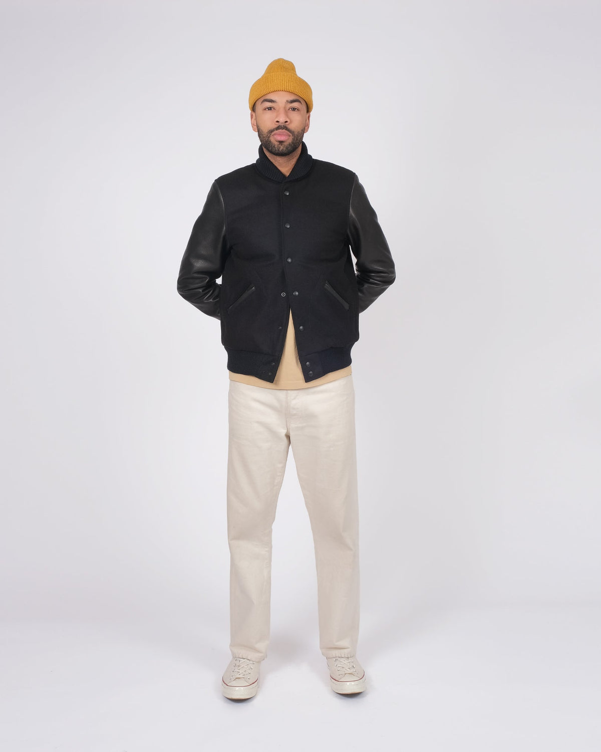 Man wearing a black leather jacket, beige pants, and a yellow beanie on a white background