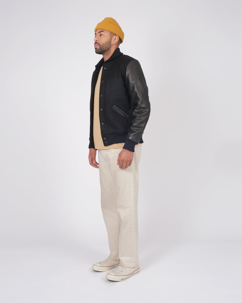 Man wearing a black leather jacket, beige beanie, and cream pants on a white background
