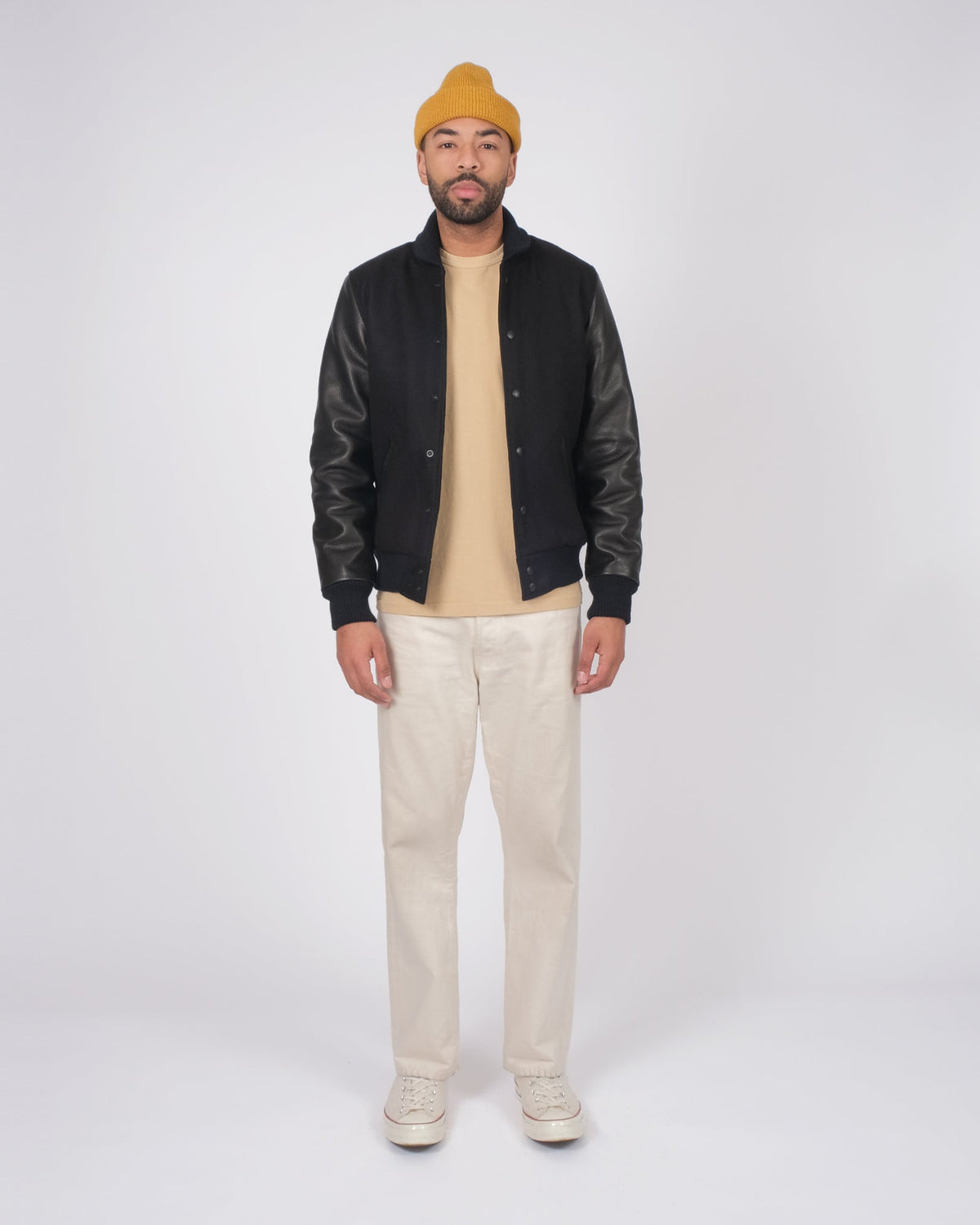 Man wearing a black leather jacket, beige sweater, cream pants, and yellow beanie on a white background