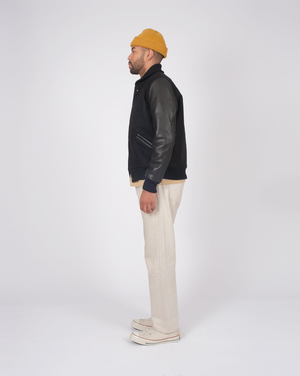 Person wearing a black leather jacket, beige beanie, and light-colored pants on a white background
