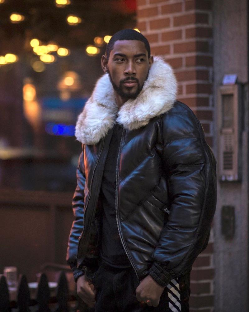 Man wearing a black leather jacket with a white fur collar standing in an urban setting.
