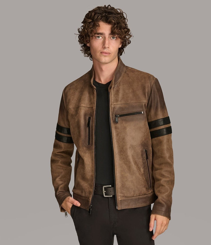 A man wearing a brown suede-textured moto jacket with black stripe details on the sleeves, styled over a black t-shirt and black pants, against a solid gray background.